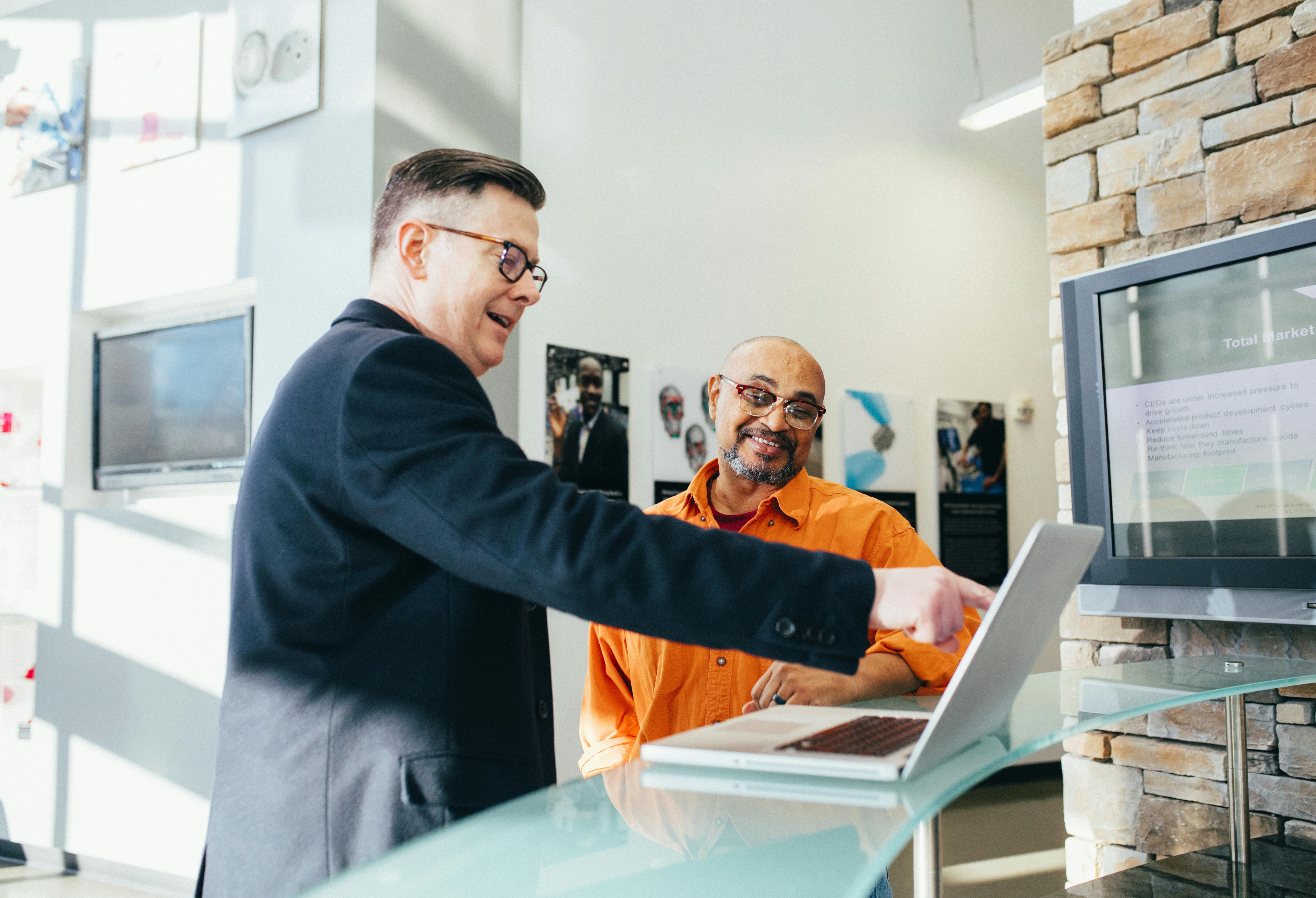 Dois homens, um de camisa preta e outro de camiseta laranja, sorriem e olham para o computador. Entenda como a consultoria de vendas é importante durante o lançamento imobiliário